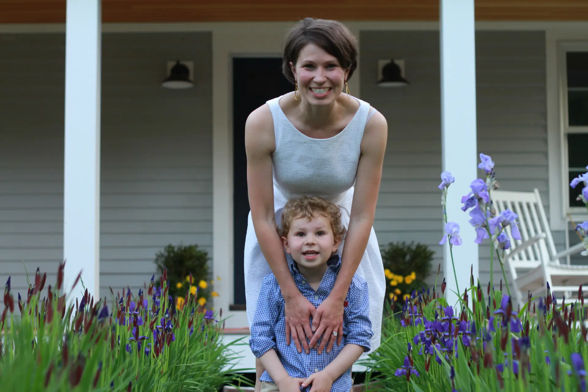 A smiling woman in a sleeveless white dress stands behind a young boy with curly hair in a blue shirt, both surrounded by blooming purple flowers in front of a house with a porch.