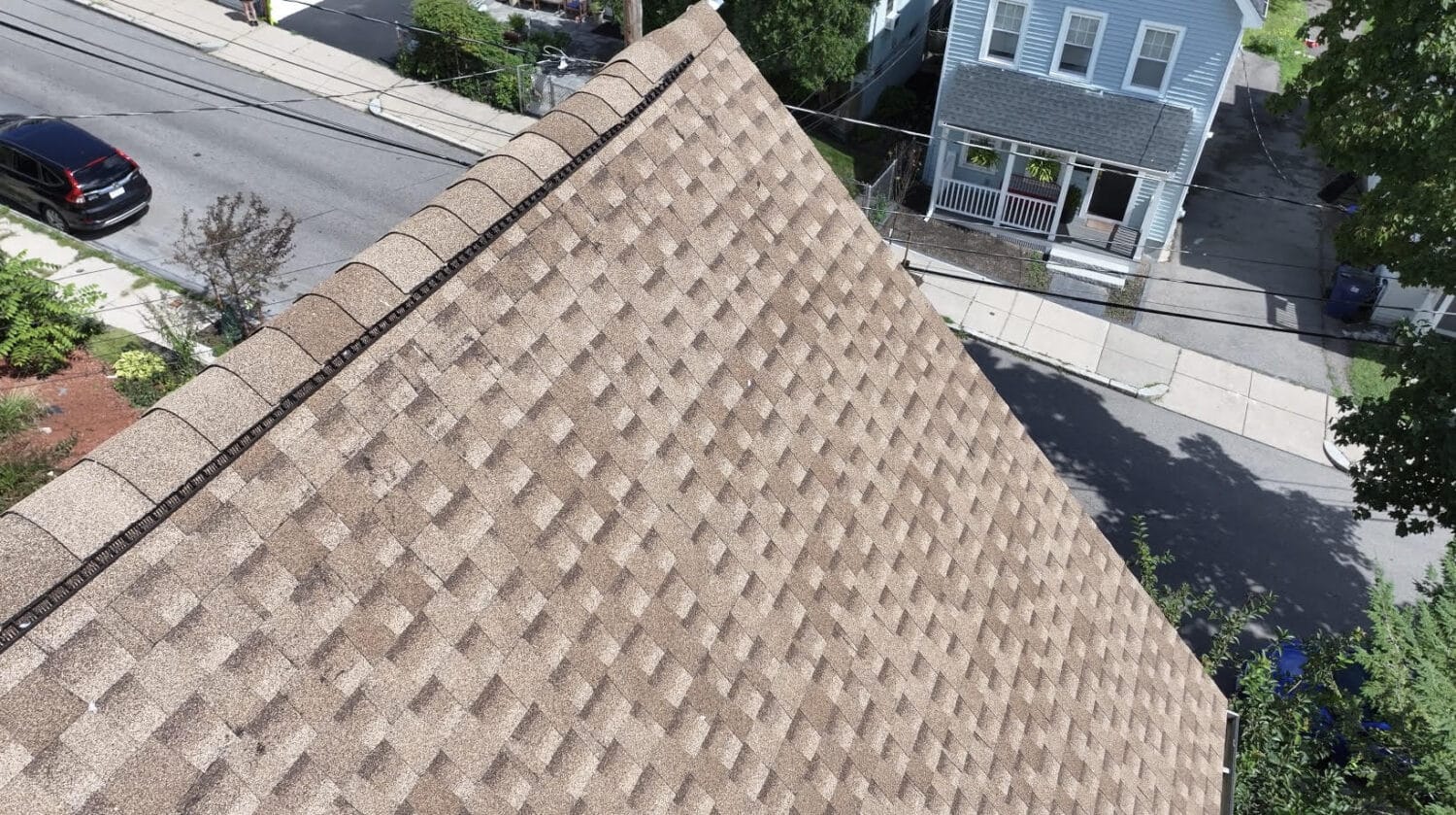Aerial view of a brown shingle roof on a house, featured in our spring roof guide, with neighboring homes, a street, and parked cars below. Trees and sidewalks line the residential area.