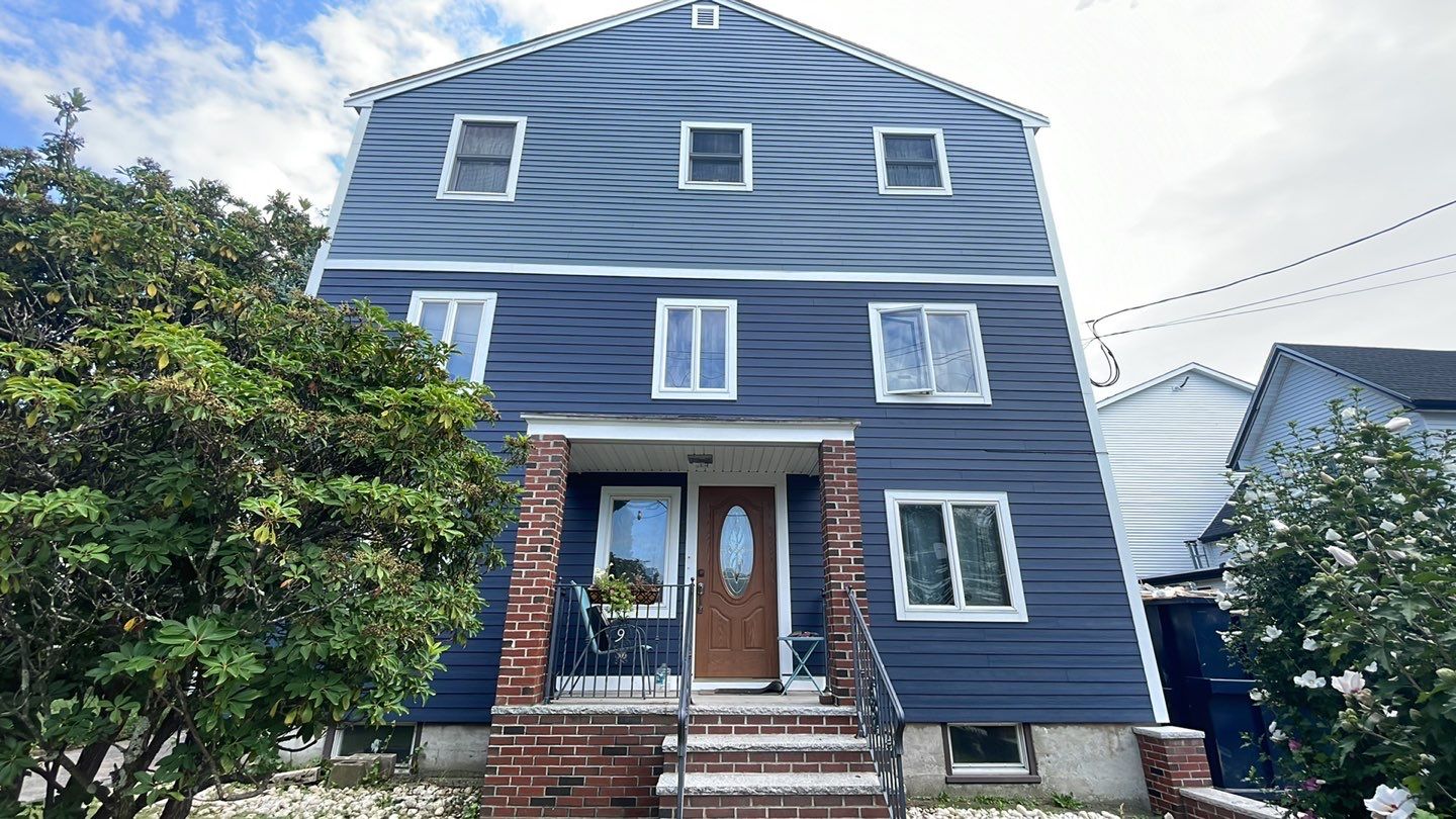 A modern, three-story blue house with a brick facade at the entrance and condensation forming on the windows. There are two front doors, each with a small porch and steps leading up. Surrounding the house are a few bushes beneath the crisp winter sky above.
.