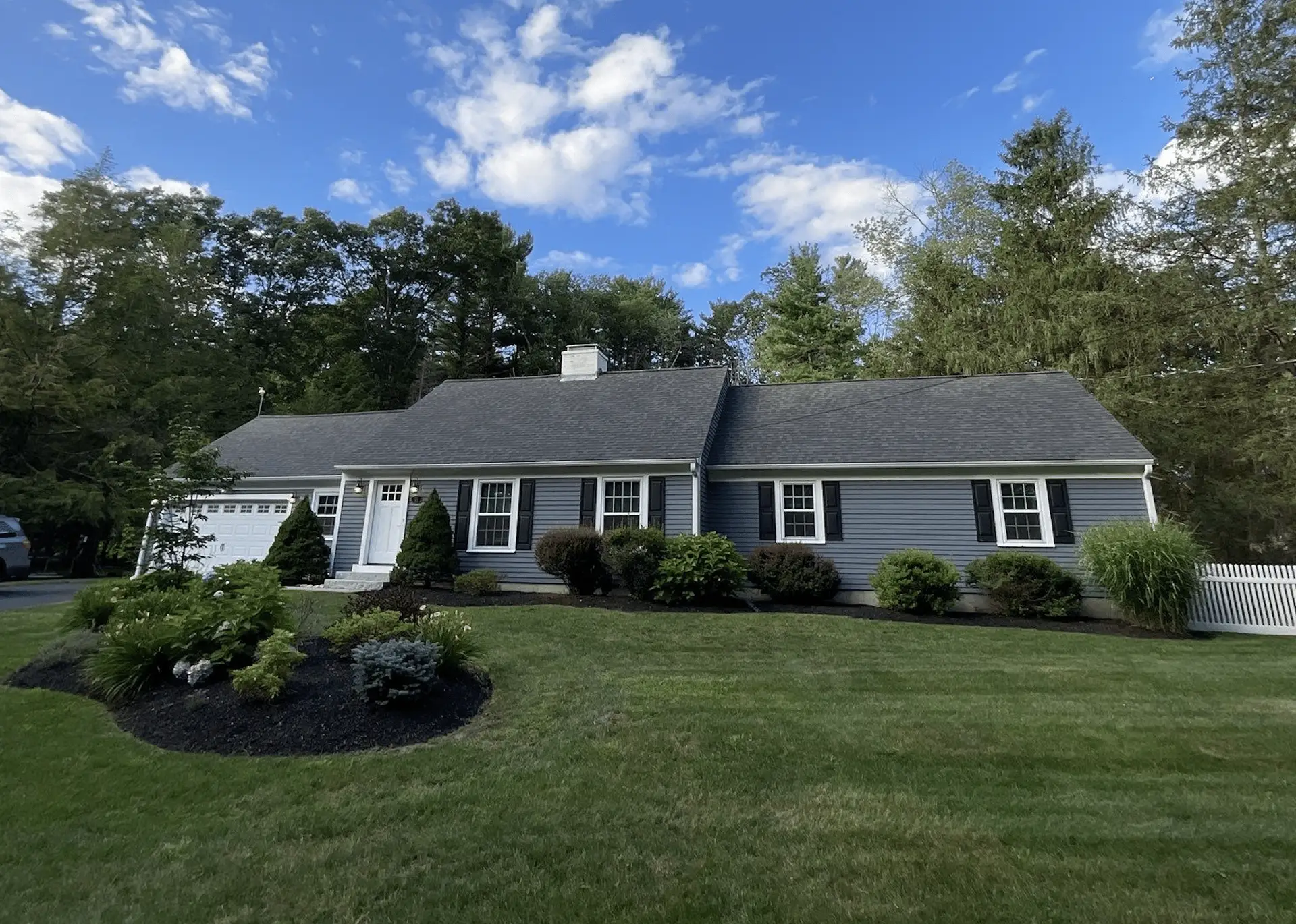 A single-story gray house with white trim and a shingled roof, designed to keep siding from fading in the sun, is surrounded by green grass, shrubs, and trees under a blue sky with scattered clouds.