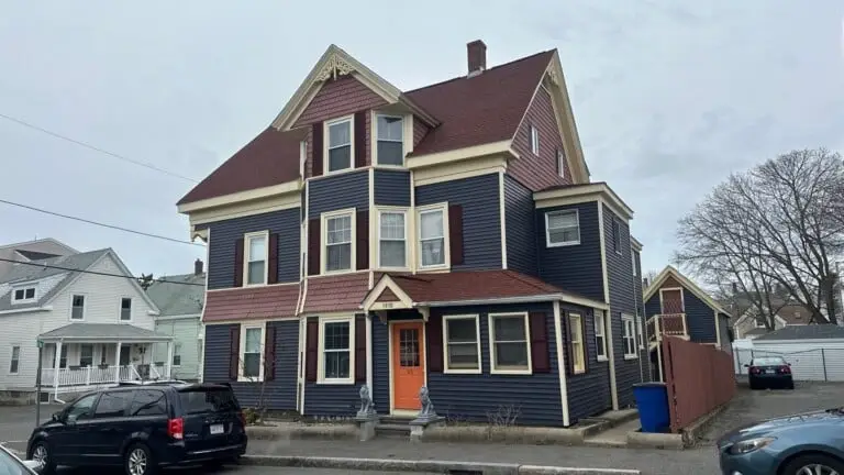 A two-story suburban house with beige siding, dark gray roof installed by a reputable roofing company in Rockport MA, and an attached double garage. The front yard has green grass, a few trees, and a driveway leading to the garage, with woods in the background. Roofing in lawrence ma