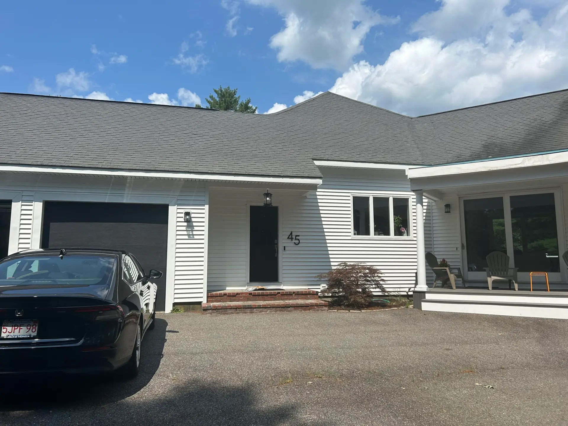 A two-story suburban house with beige siding, dark gray roof installed by a reputable roofing company in Rockport MA, and an attached double garage. The front yard has green grass, a few trees, and a driveway leading to the garage, with woods in the background. Roofing in Tyngsborough MA