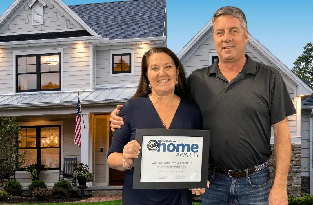 A smiling woman and man stand in front of a modern house. The woman holds a framed Northshore Home Awards certificate, celebrating Coastal Windows & Exteriors Wins North Shore Magazines BONS 2024 Award. An American flag waves near the entrance.