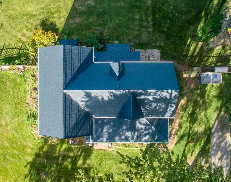 A two-story suburban house with beige siding, dark gray roof installed by a reputable roofing company in Rockport MA, and an attached double garage. The front yard has green grass, a few trees, and a driveway leading to the garage, with woods in the background. Roofing in lawrence ma