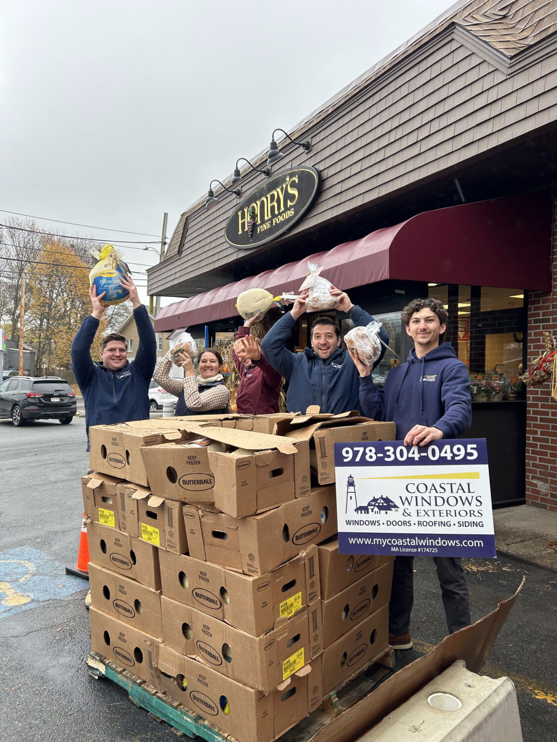 Five people stand outside Henrys Fine Foods, smiling and holding bags of donated bread above their heads, beside a large stack of boxes and a “Coastal Windows & Exteriors” sign, showcasing “Freedom from Want” with Turkey Donation.