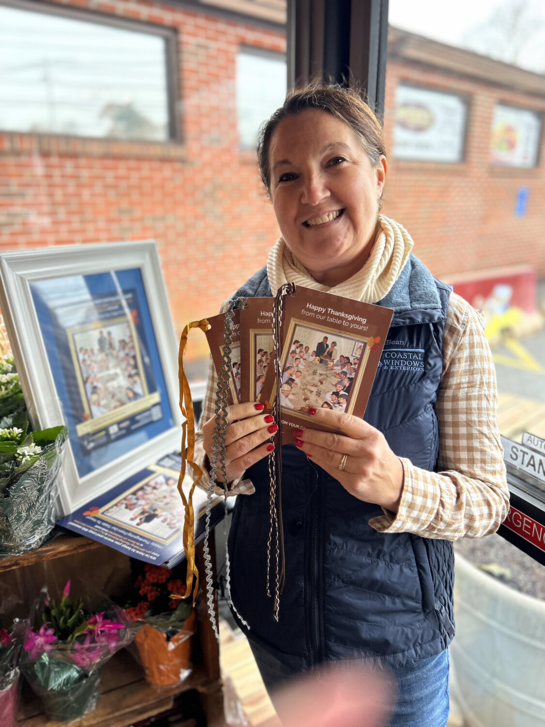 A smiling woman stands indoors by a window, holding several Thanksgiving-themed booklets. Showcasing “Freedom from Want” with Turkey Donation, she wears a checked shirt with a navy vest beside a display of framed pictures and colorful flowers.