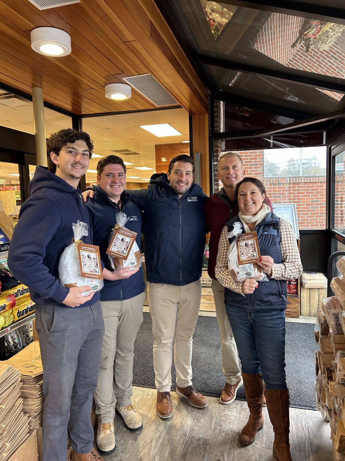 Four men and one woman stand together inside a store entrance, smiling and holding packaged baked bread, as the group showcases “Freedom from Want” with Turkey Donation. Cardboard boxes are stacked nearby, with shelves and windows in the background.