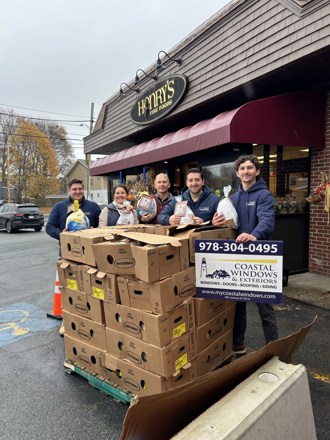 Five people stand outside Henrys Fine Foods, smiling and holding bags of food behind a large stack of boxes. A sign for Coastal Windows & Exteriors is propped against the boxes, showcasing “Freedom from Want” with a Turkey Donation.