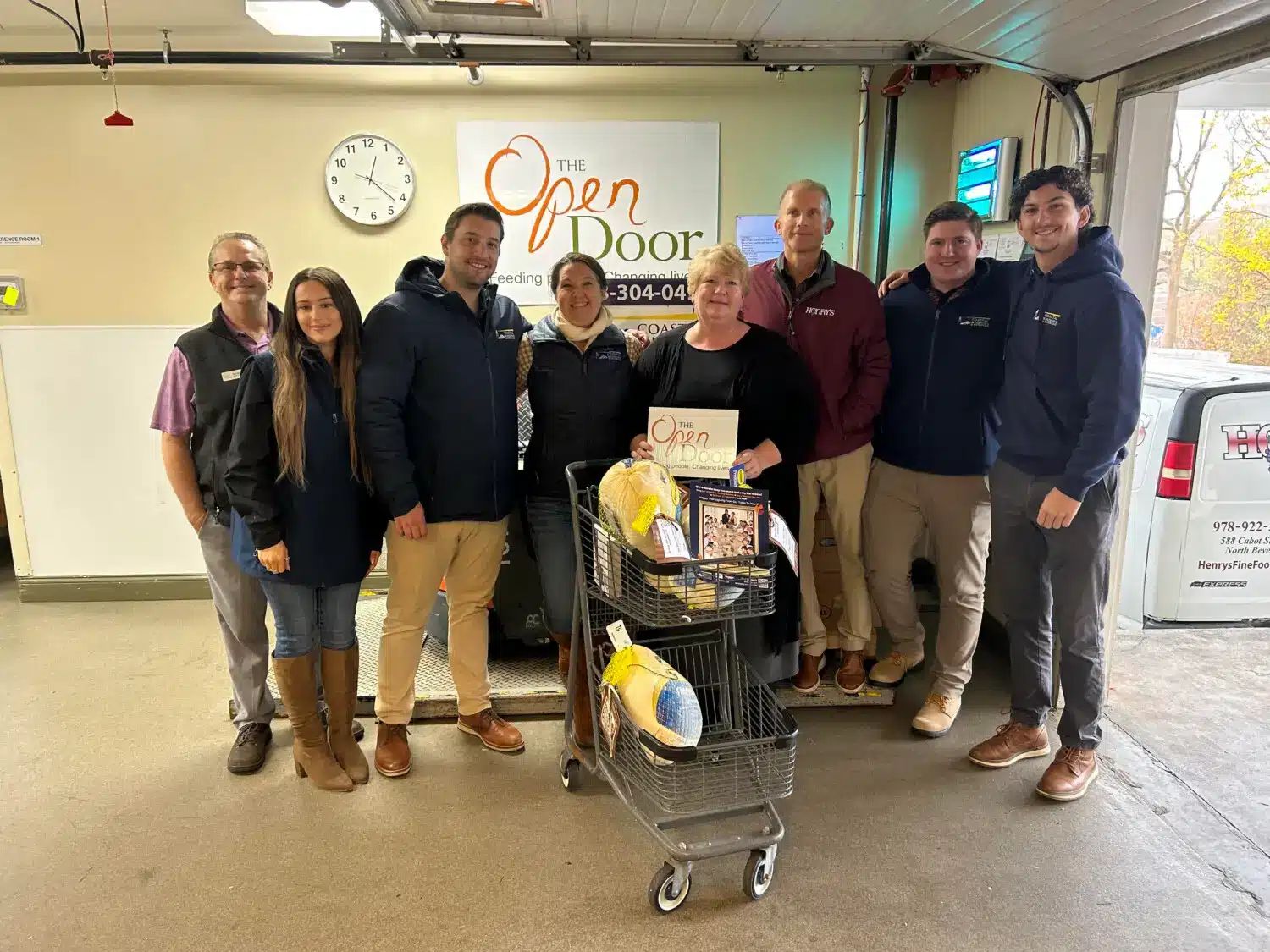 Eight people stand indoors, smiling around a shopping cart filled with turkeys and food items, showcasing “Freedom from Want” with Turkey Donation for The Open Door food pantry. A clock and The Open Door logo are visible in the background.