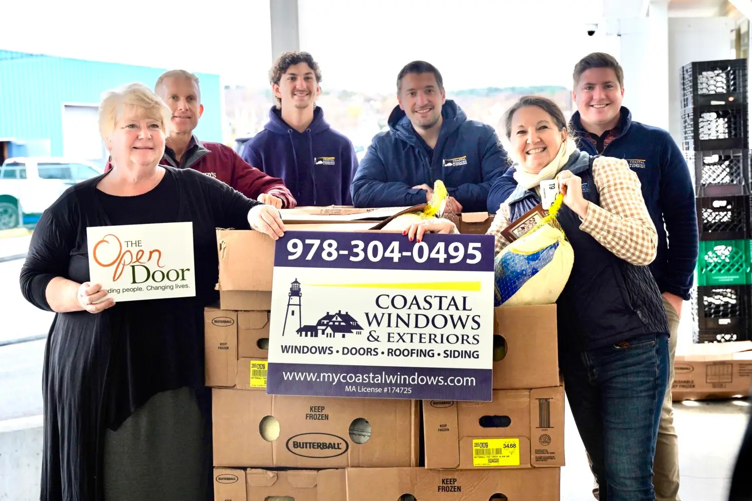 Six smiling people stand behind boxes, holding a “Coastal Windows & Exteriors” sign and a “The Open Door” sign, showcasing “Freedom from Want” with Turkey Donation; crates and a building are in the background, highlighting their collaborative effort.