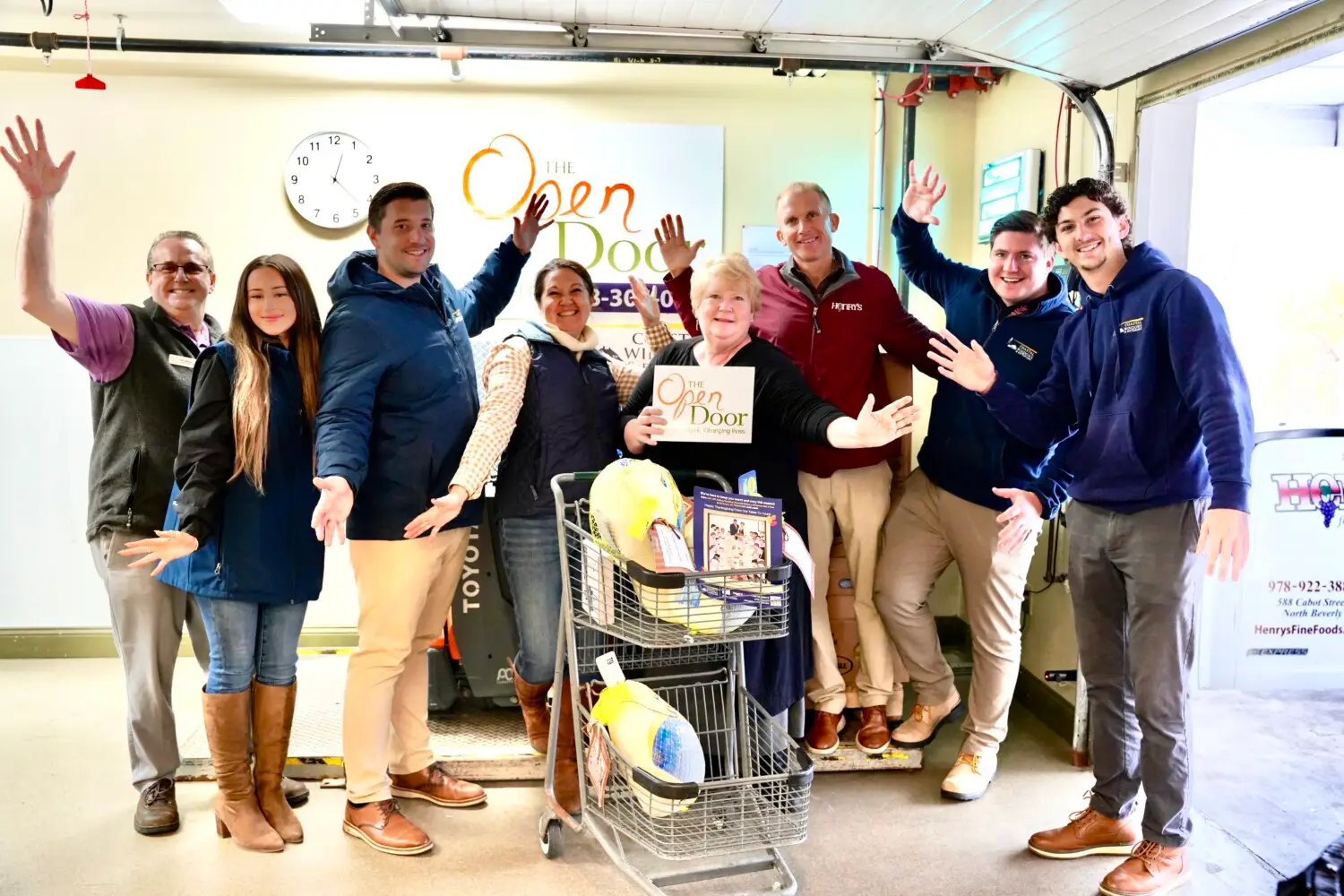 A group of nine smiling people stand around a cart filled with turkeys and food at The Open Door food pantry, posing with raised arms in a garage-like setting under a wall clock, showcasing “Freedom from Want” with their turkey donation.