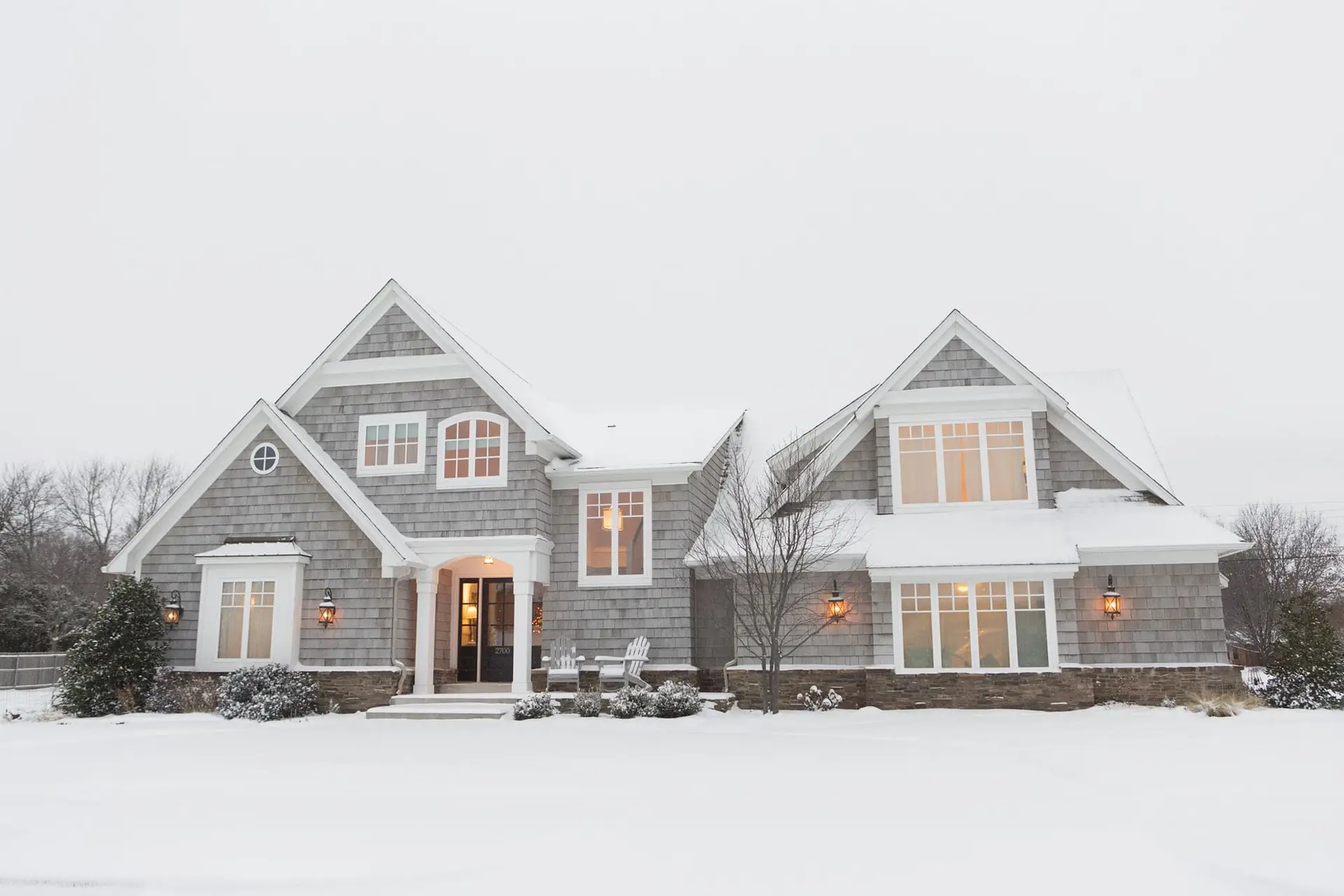 A large, two-story gray house with peaked roofs and big windows is surrounded by snow. Warm lights glow inside as Coastal Windows & Exteriors reminds homeowners to protect your home this winter with a sound roof for roofing readiness.