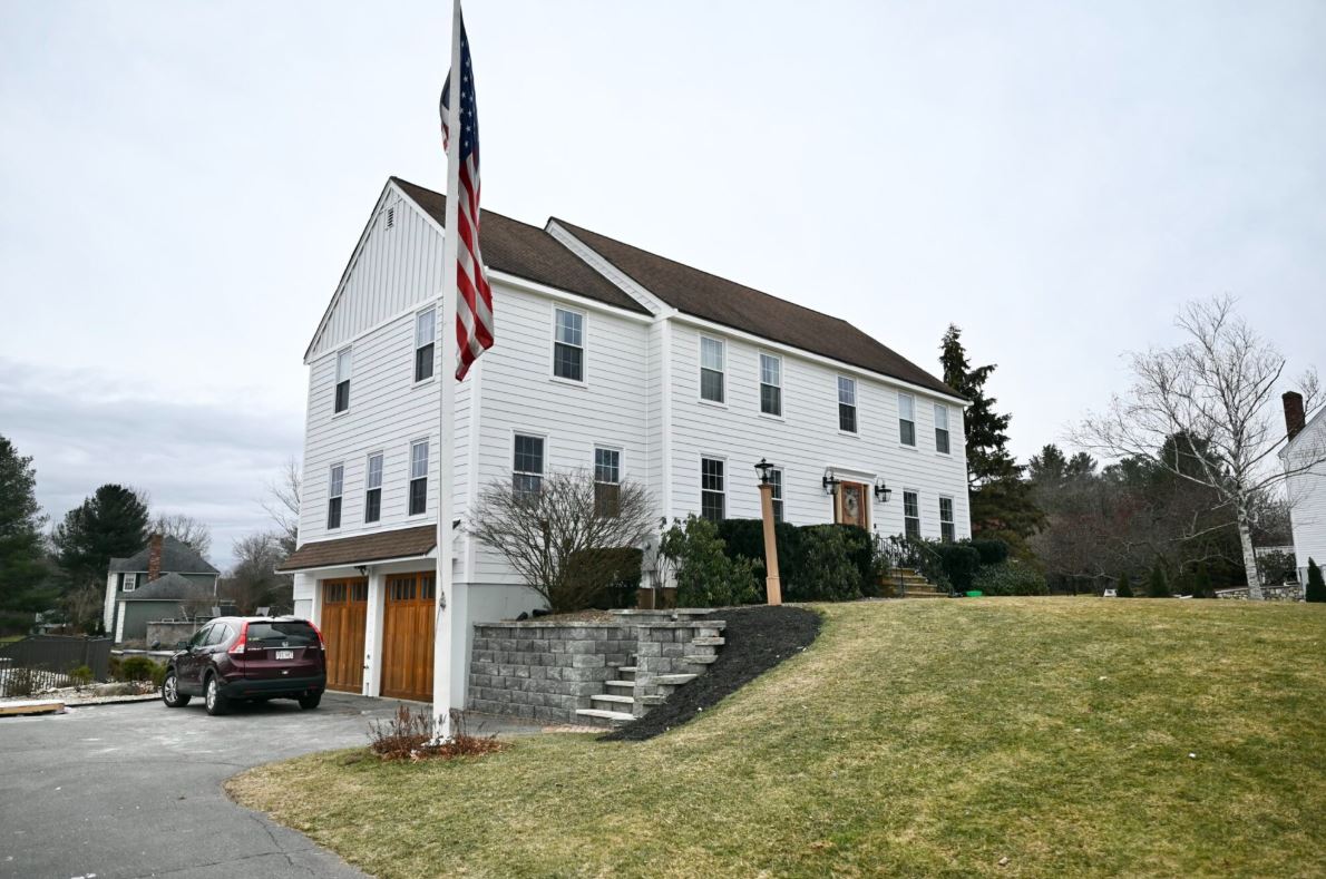 This two-story white house in North Andover features a brown roof, Colonial Makeover Siding, attached garage, raised front yard, and an American flag by the driveway. A car is parked nearby with trees in the background under an overcast sky.