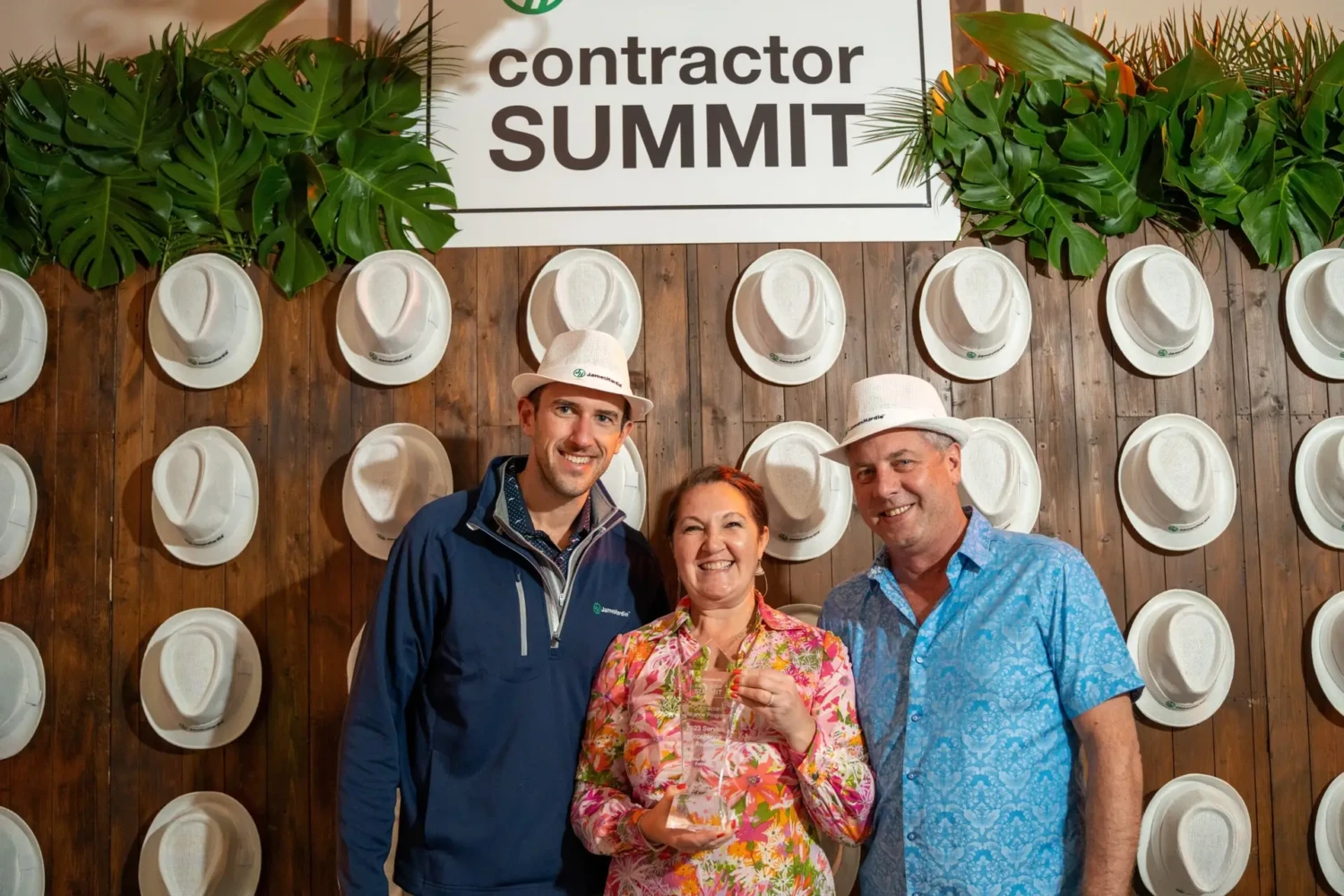 Three people stand smiling in front of a wall decorated with white hats under a contractor SUMMIT sign. The woman in the center holds a james hardie service excellence trophy, and all wear white hats. Lush green leaves frame the sign above.