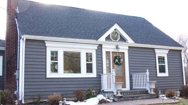 A small, single-story Classic Cape house with dark gray siding and white trim features a steeply pitched roof and a brick chimney. The entrance, accented by two white planters, has a wooden door flanked by windows. A few steps lead up through the snow-covered ground.