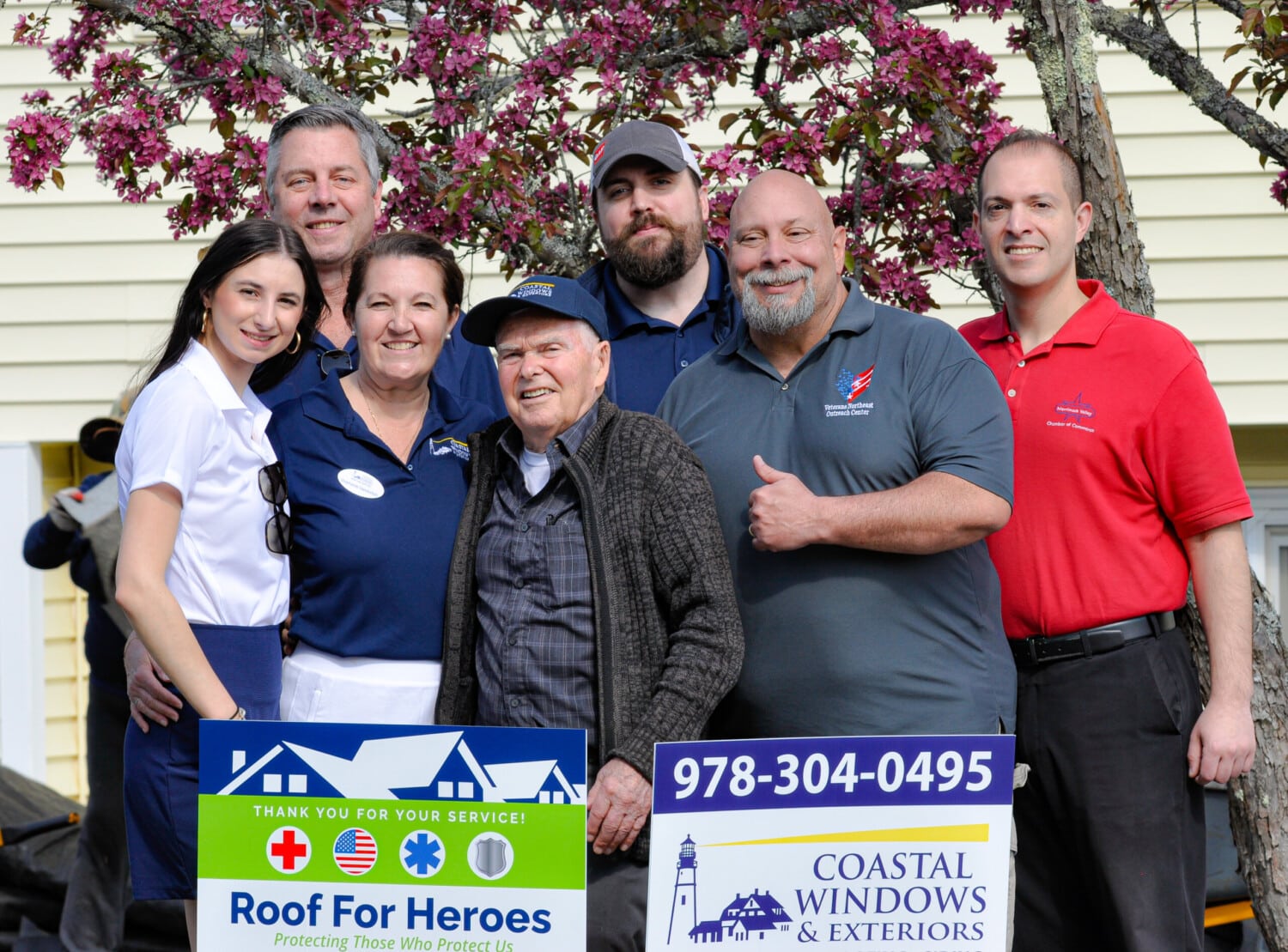 A group of seven people, including an elderly man, smile outdoors in front of a flowering tree and house, standing behind signs for Roofs for Heroes and Coastal Windows & Exteriors as they celebrate the installation of a new GAF roof.