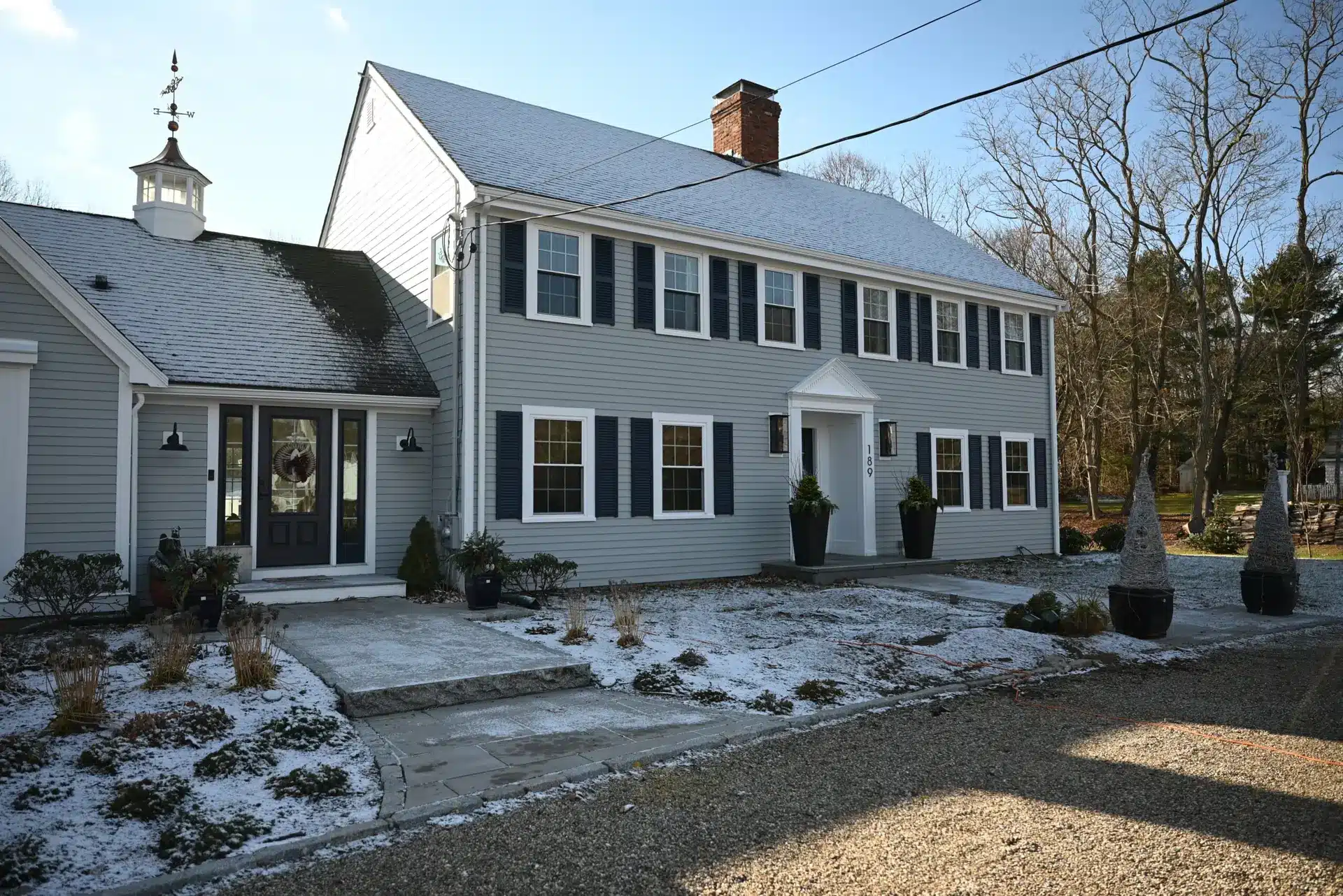 A two-story gray colonial-style house with black shutters, a white front door, and light snow covering the ground and roof—showing how winter energy bill concerns rise as cozy homes brace against the chill. Plants sit by the entry; leafless trees frame the background.