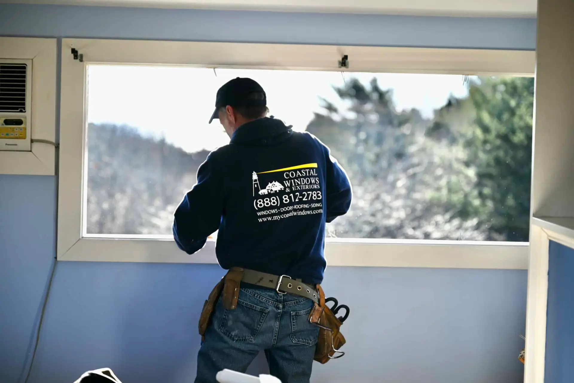 A worker wearing a tool belt and a Coastal Windows & Exteriors sweatshirt performs window installation in MA, repairing or installing a window in a light blue room with trees visible outside.