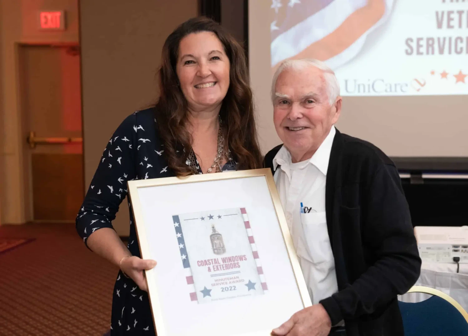 A woman and an older man smile while holding a framed certificate together at an indoor event, celebrating Coastal Windows & Exteriors Chosen as Honoree for the 2022 Minuteman Service Award. A projection screen and chairs are visible in the background.