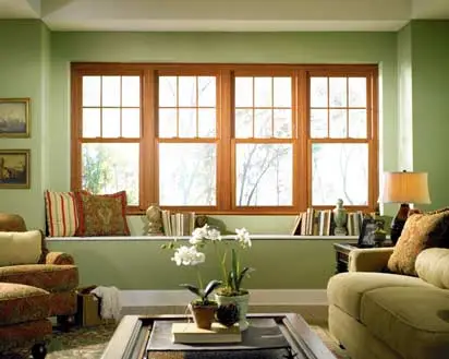 A cozy living room with green walls, large wooden-framed windows showcasing elegant window styles, beige and patterned sofas, decorative pillows, a table with potted plants, books on the windowsill, and a table lamp.