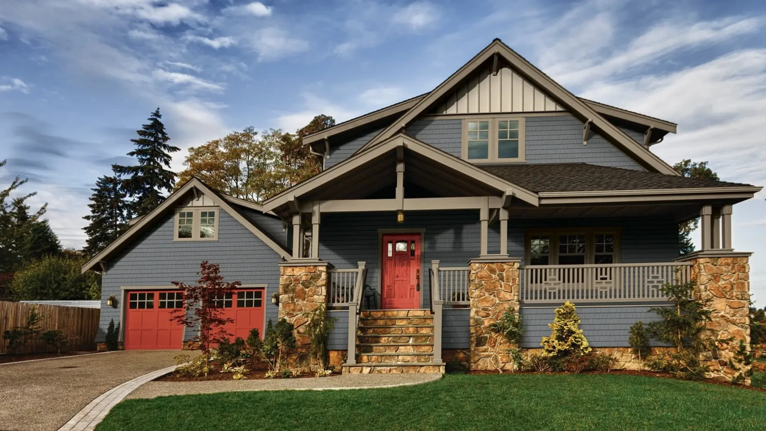 Two-story blue house with board and batten vinyl siding, stone accents, a covered front porch, red front door, and attached garage with red doors, surrounded by greenery under a partly cloudy sky.