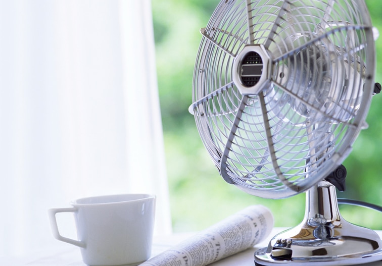 A silver tabletop fan sits next to a white coffee cup and a rolled newspaper on a bright windowsill with a lush green background outside—perfect for those who want to stay cool without air conditioning.
