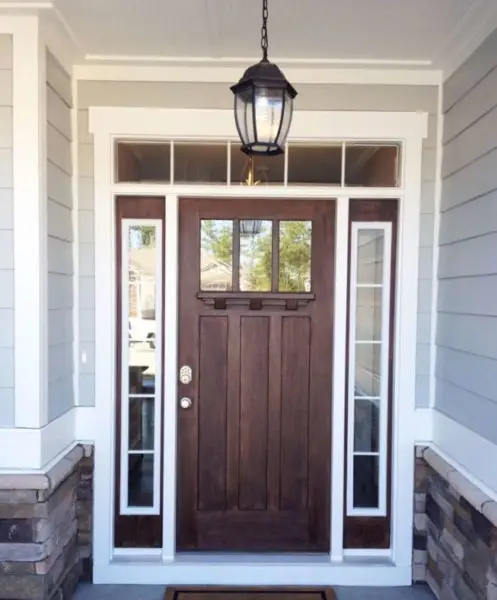 A wooden front entry door with a small glass window at the top, flanked by vertical glass sidelights and a rectangular transom window above. The exterior features light blue siding and stone accents. A black lantern hangs overhead, highlighting classic entry door options.