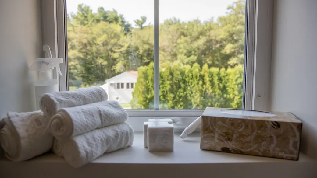 Rolled white towels, a tissue box, a spray bottle, and a small container sit on a windowsill—perfect when choosing windows that showcase lovely views of trees and a house outside.