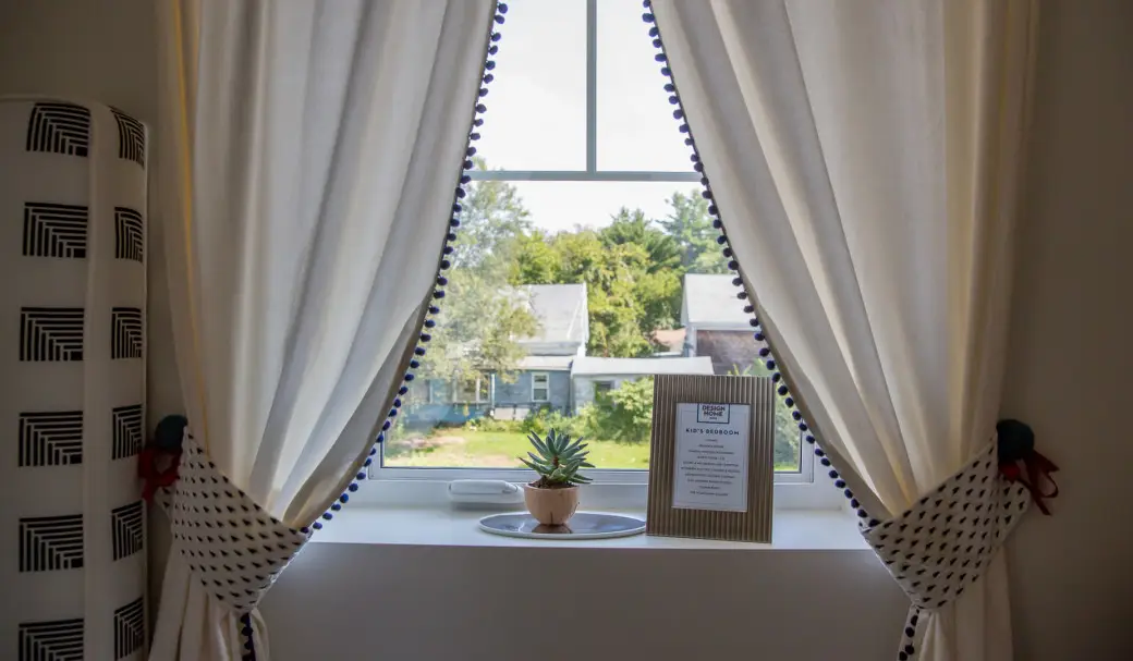 A window framed by white curtains with blue trim, tied back to reveal a sparkling view thanks to recent window cleaning. On the windowsill are a small potted plant and a framed notice or sign, with houses and trees visible outside.