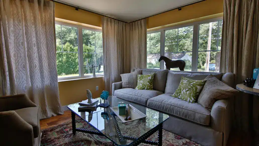 A cozy, energy efficient living room with large windows, beige curtains, a light gray sofa with green patterned pillows, a glass coffee table, an armchair, and a stylish horse figurine on the windowsill.
