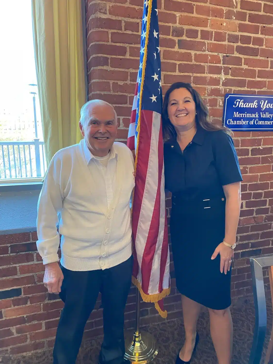 Two people stand smiling beside an American flag indoors, in front of a brick wall. The man on the left wears a white sweater, while the woman on the right wears a dark dress. A Thank You sign highlights their FREE ROOF Donation to Local Veteran.