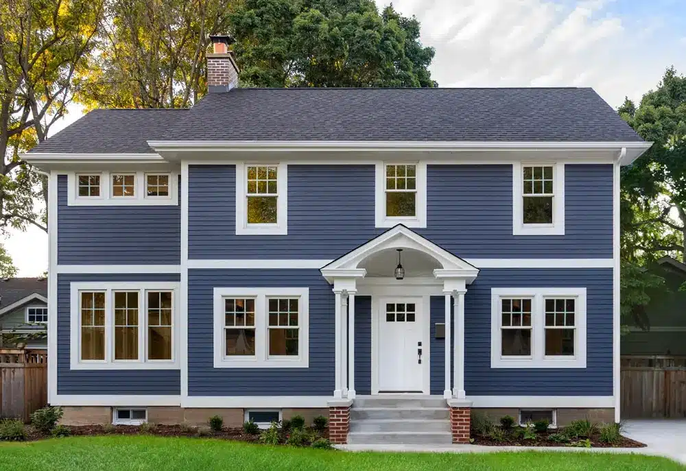 A two-story house with blue siding and white trim. It features a symmetrical facade with six windows and a centered front door framed by white columns. The house sits on a grassy lawn with trees in the background.