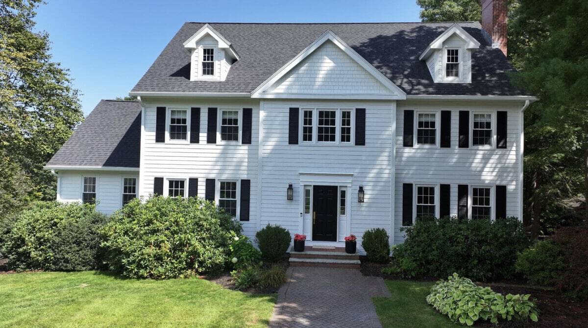 A white two-story house with dark shutters and dormer windows. A brick path leads to the front door, flanked by two small shrubs in pots. Lush green bushes and grass surround the house under a clear blue sky.