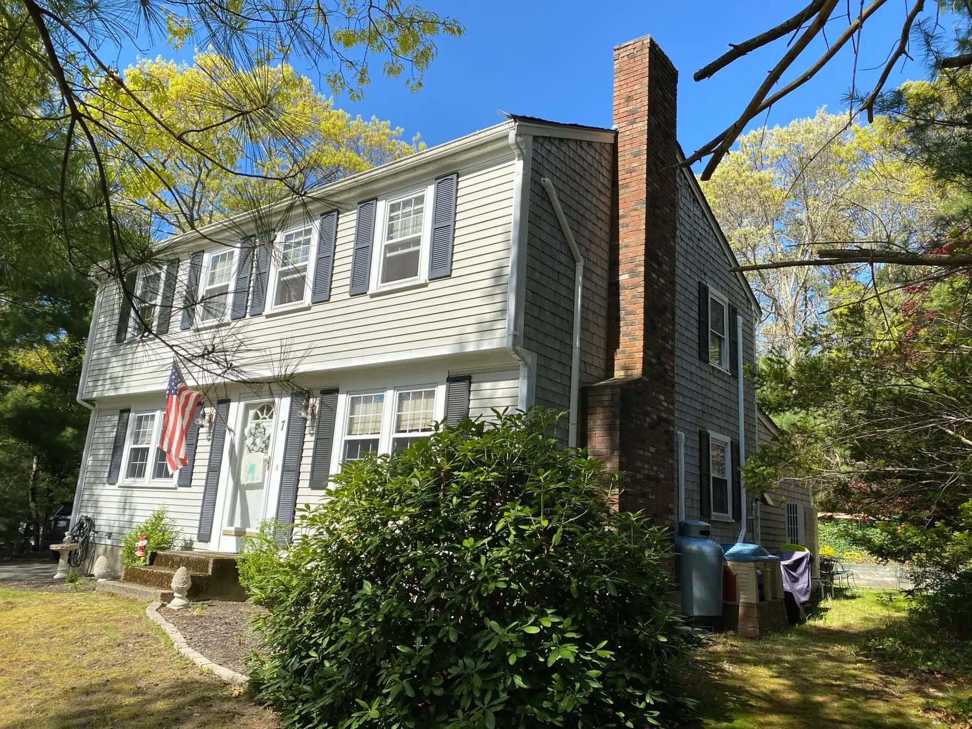 A two-story gray house with white trim, black shutters, and vinyl siding—ideal if you’ve wondered, “Can I paint vinyl siding?”—features a brick chimney and an American flag by the front door, framed by lush shrubs under a clear blue sky.