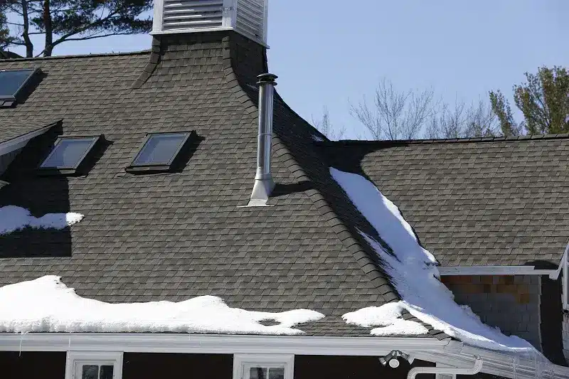 A house with a steep, shingled roof partially covered in melting snow hints at the challenges to replace a roof in winter. Skylight windows, a chimney, and vent stand out, with trees and a clear blue sky in the background.