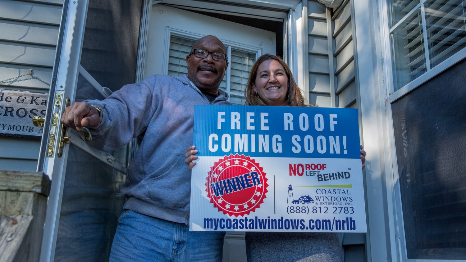 A smiling man and woman stand in a doorway, floored by a free roof, holding a blue sign that says FREE ROOF COMING SOON! WINNER, with contact info for Coastal Windows & Exteriors and the website ppc.mycoastalwindows.com/nrlb.