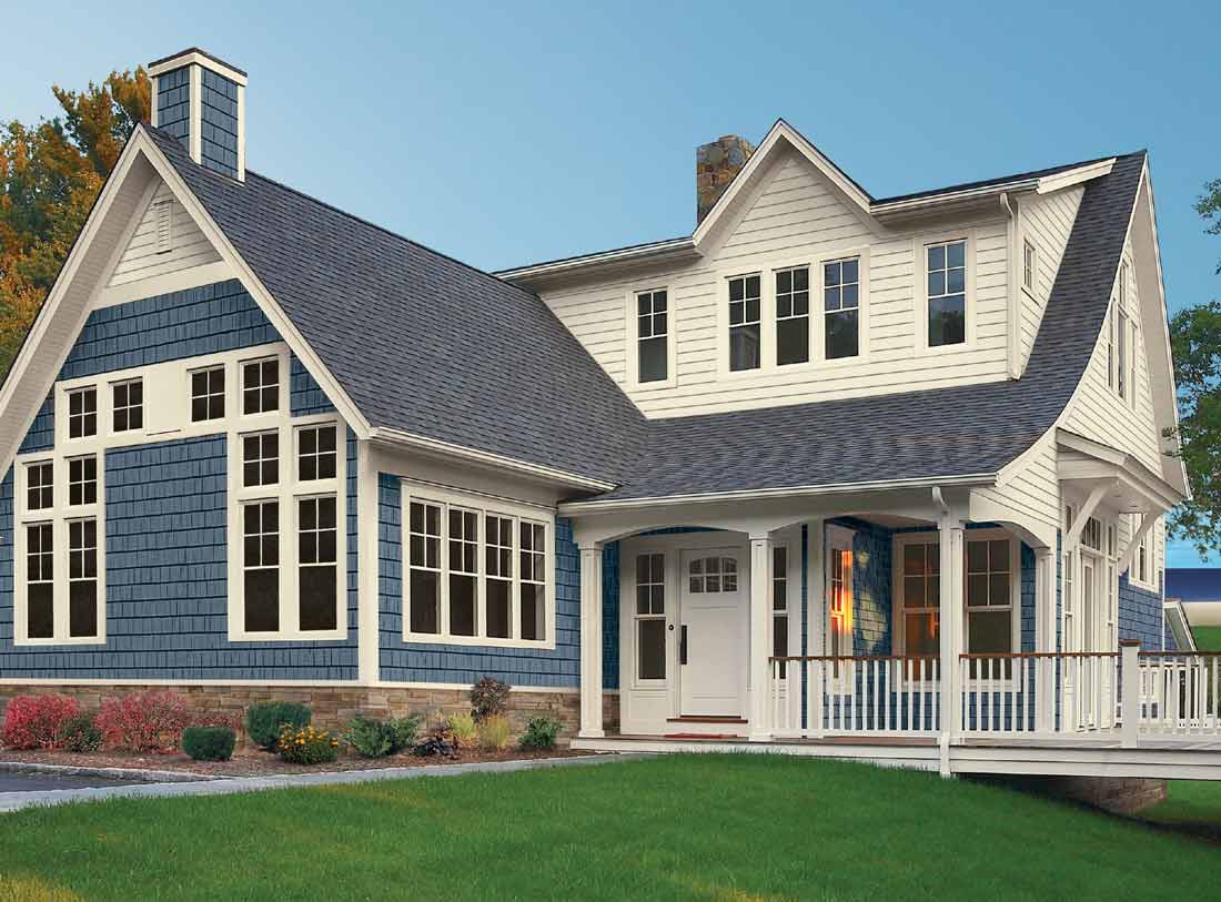 A two-story house with a large front porch exudes charm, boasting a mix of blue and white siding and energy-zapping windows. The dark gray roof complements the stone chimney. The neatly landscaped yard, with its green grass and shrubs, enhances this inviting homes allure.