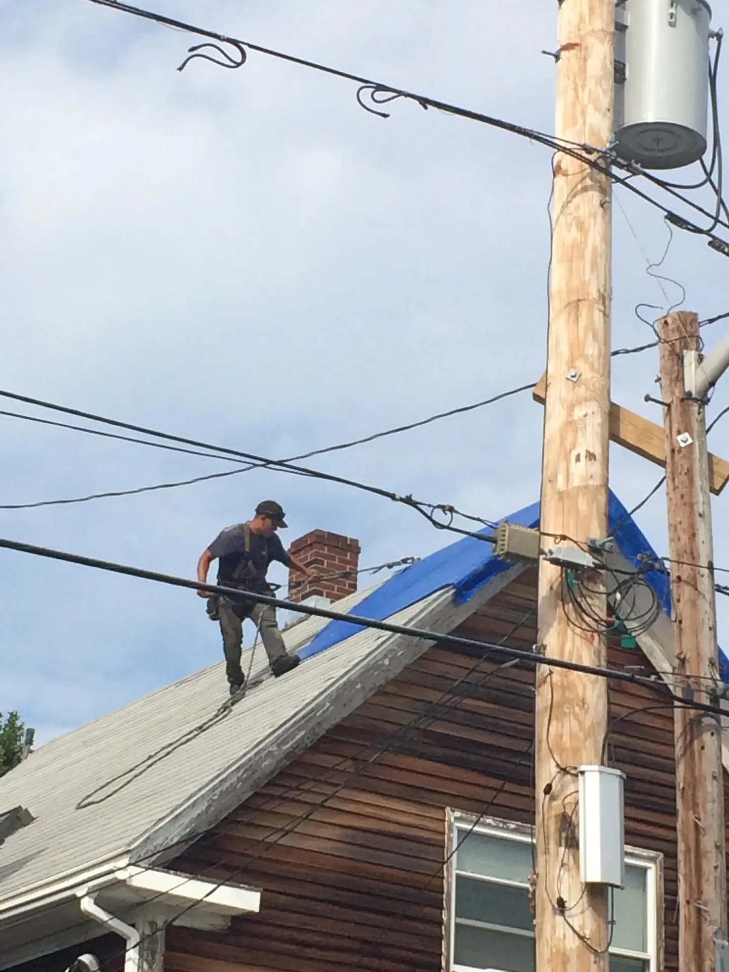 A man stands on the sloped roof of a wooden house, painting part of it blue as part of the Revere Roof Rescue program. Power lines and utility poles stretch across the cloudy sky in the foreground.