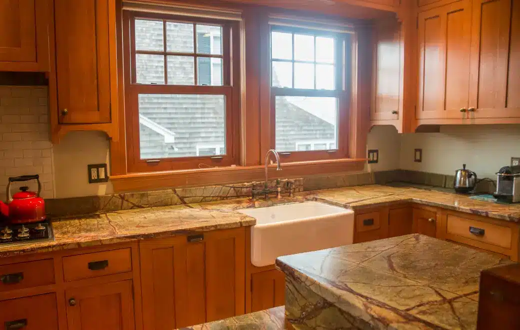 A cozy kitchen with wooden cabinets and marble countertops features a farmhouse sink centered under two large windows. A red kettle sits on the stove, and a toaster is on the right. Without air conditioning, the windows offer a spot to cool off while enjoying a view of rooftops.