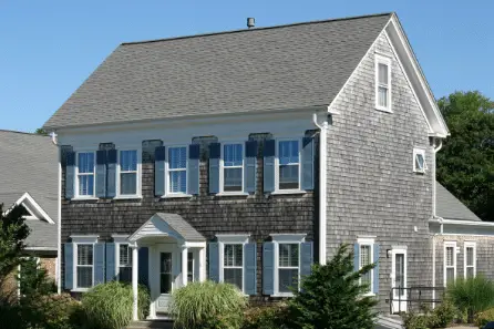 Two-story house with weathered gray shingle siding, white trim, blue shutters, and a small front porch—classic elements inspired by remodeling trends in New England. Tall grasses and shrubbery accent the front beneath a clear blue sky.