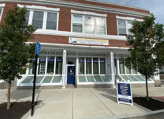 A brick building with a sign reading Coastal Windows & Exteriors above the blue door; windows display service offerings. If you’re hiring a contractor, check out the sandwich board and blue handicap parking sign by the small trees out front.