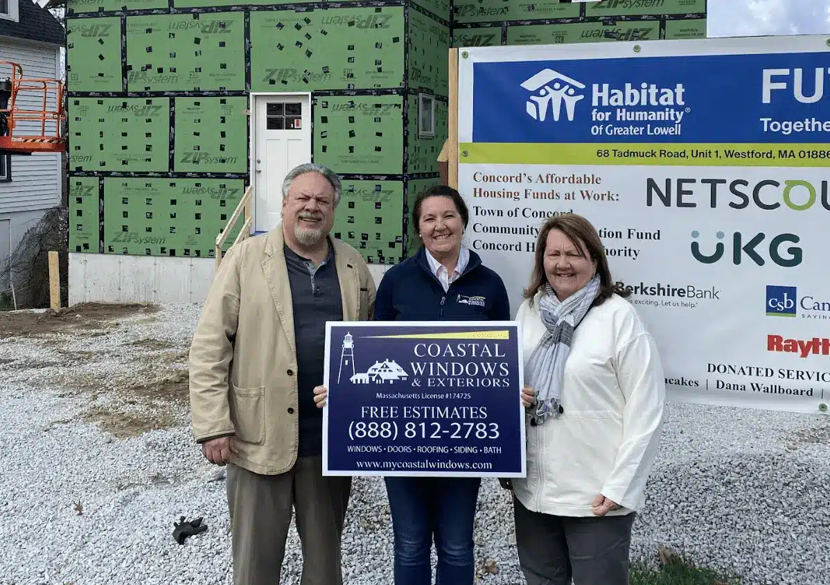 Three people stand smiling in front of a house under construction in Concord MA, with a Habitat For Humanity sign. The person in the middle holds a Coastal Windows & Exteriors sign offering free estimates.