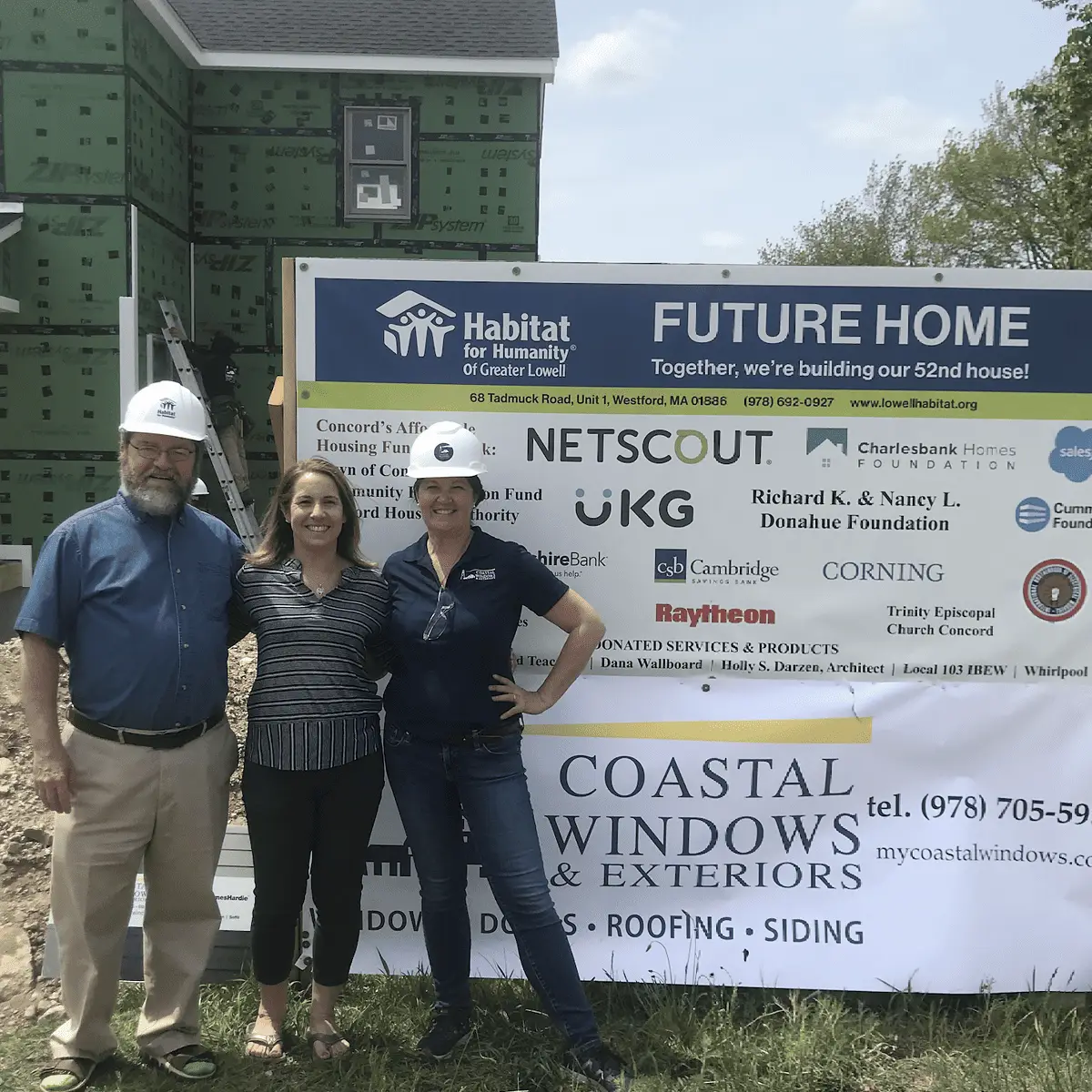 Three people in hard hats smile in front of a Habitat for Humanity “Future Home” sign and a partially constructed house in Concord MA. The sign displays sponsor logos and contact info for Coastal Windows & Exteriors.