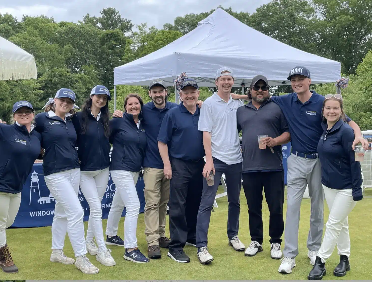 A group of nine people pose and smile together outdoors on grass in front of a white tent at the Northshore Scramble Charity Golf Tournament. Most wear navy shirts, white pants, and caps, while two are in white; trees and a cloudy sky form the backdrop.
