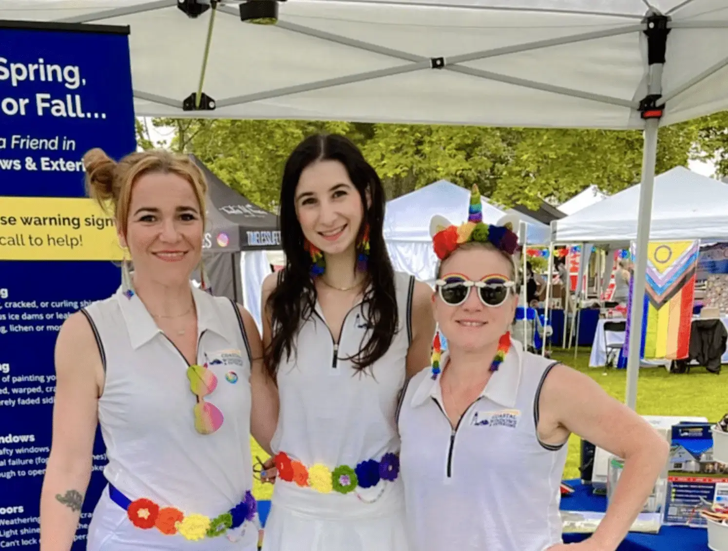 Three people stand smiling under a white canopy at North Shore Pride, wearing white outfits with rainbow-themed accessories. Colorful booths and trees in the background highlight this vibrant celebration of individuality and diversity.