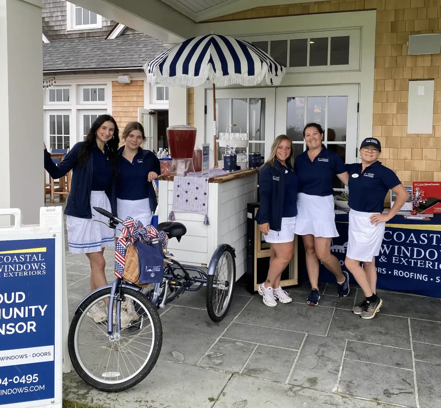 Five women in matching navy blue shirts and white skirts smile by a promotional booth under a striped canopy at the 2023 Marblehead Open Golf Tournament, with a bike in front and Coastal Windows & Exteriors signs displayed nearby.