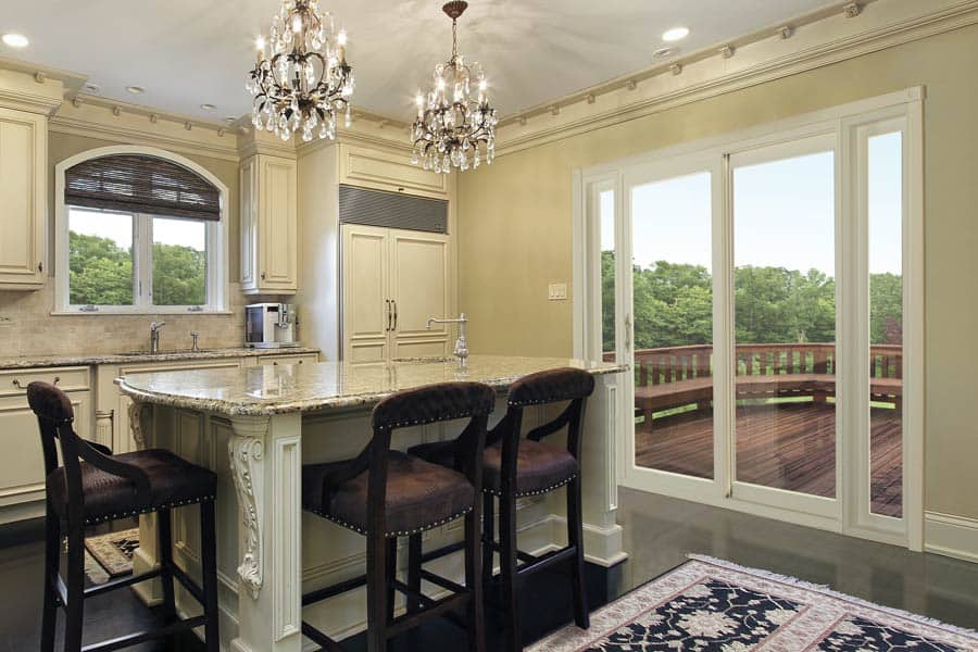 Kitchen with a large central island featuring three brown upholstered chairs. Two ornate chandeliers hang above the island. White cabinets surround the space, and sliding glass doors lead to an outdoor deck overlooking greenery. A rug lies on the floor.