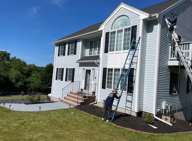 Two people work on the exterior of a two-story light gray house with vinyl siding in Woburn; one is on a ladder cleaning or painting near the roof, while the other works near the ground-level windows on a sunny day.