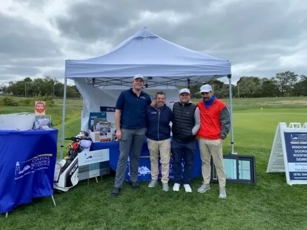 Four men stand smiling in front of a white canopy tent at the YMCA of the North Shore 2022 Glen Golf Event. Promotional materials and signs are displayed on tables around them under a cloudy sky.