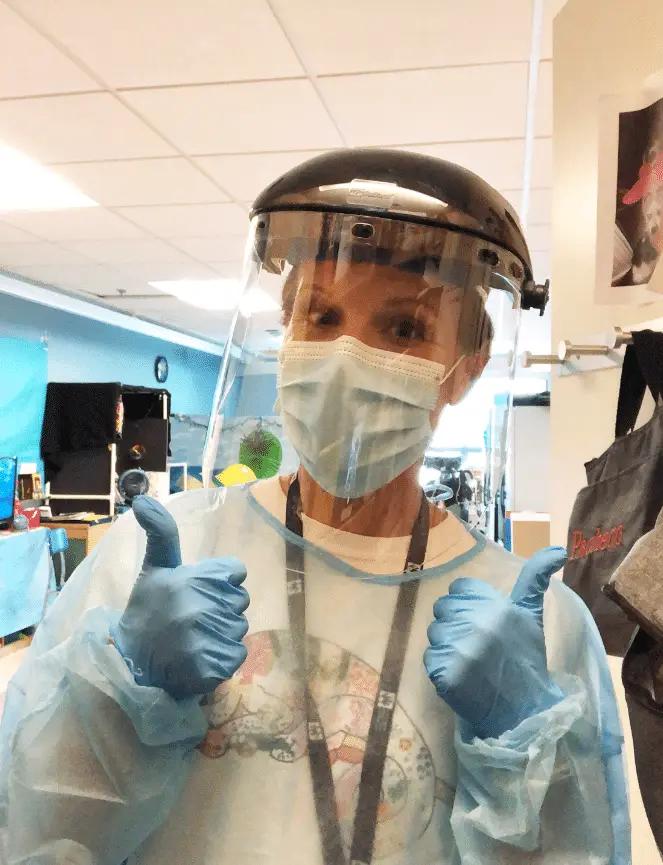 A person wearing a face shield, surgical mask, gloves, and gown gives two thumbs up in a brightly lit indoor setting, possibly a hospital or clinic—proudly recognized as roof for heroes recipient #1.
